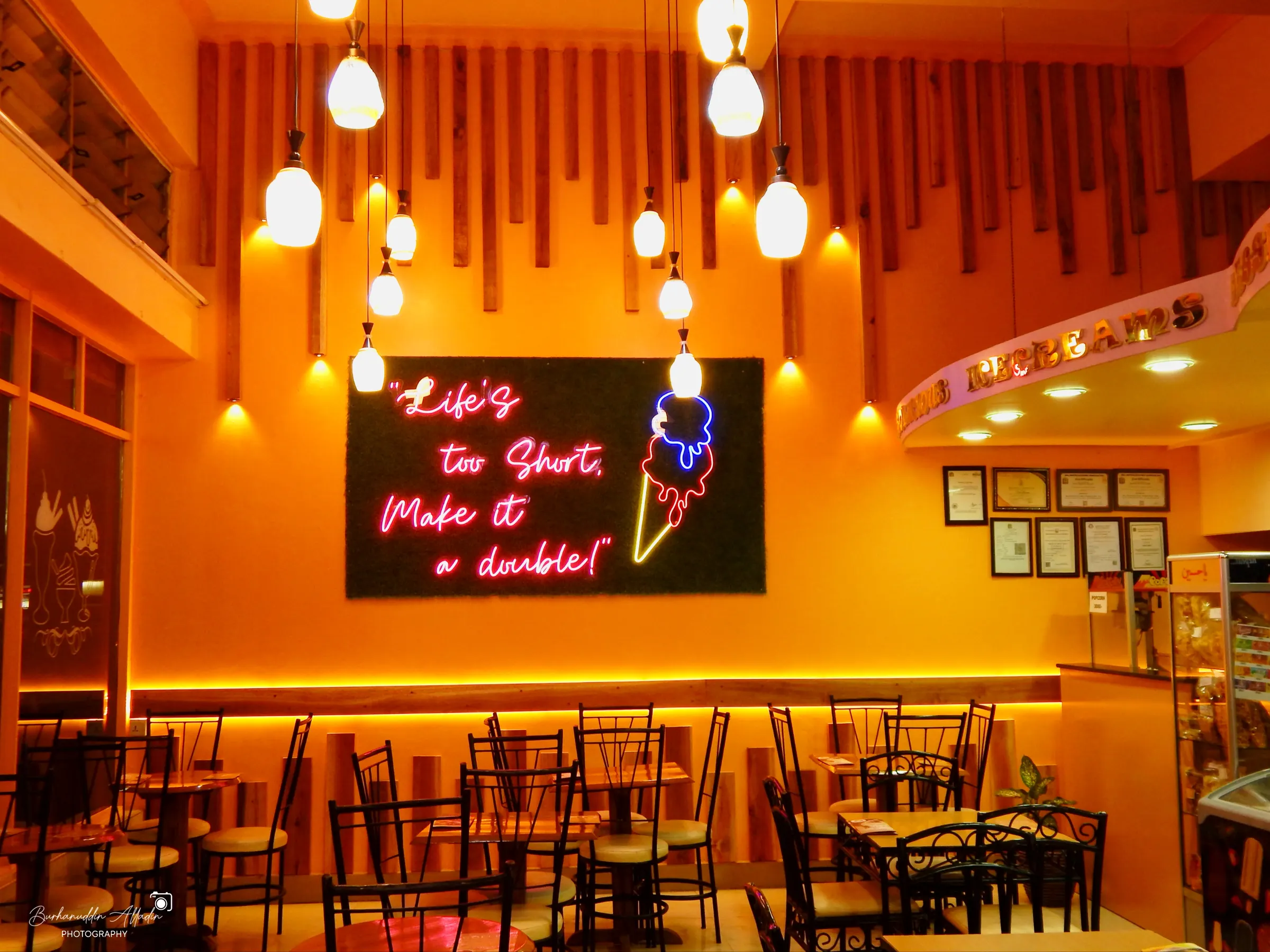 The parlour today: warm orange walls, timber slats, pendant lights, and a pink neon sign reading “Life's too short, make it a double!”